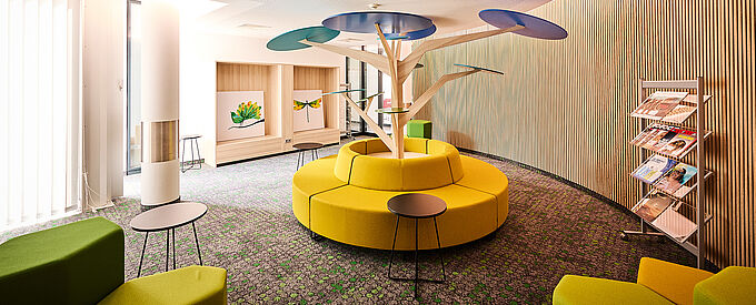 Kollaborationsbereich mit farbenfrohen Sitzpolstern und einem minimalistischen Deko-Baum in der Raummitte