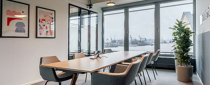 Moderner Besprechungsraum mit großem Holztisch und gepolsterten Stühlen, bodentiefen Fenstern mit Blick auf den Hamburger Hafen, Glaswänden, Wandgrafiken und einer Pflanze als grüner Akzent.